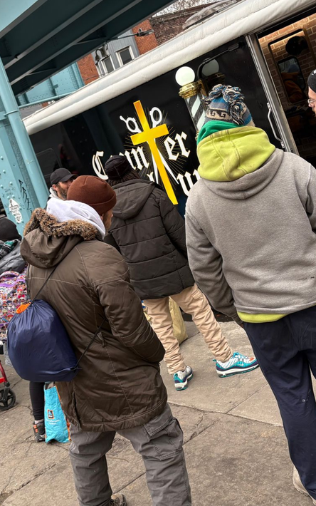A group of people wearing winter jackets stand in a line outside a mobile barber shop with a yellow cross logo on the side.