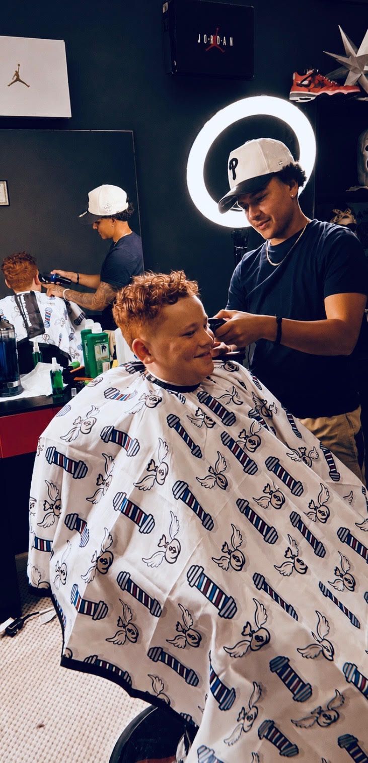 A person sits in a barbershop chair, wearing a light blue cap, denim jacket, jeans, and patterned sneakers.