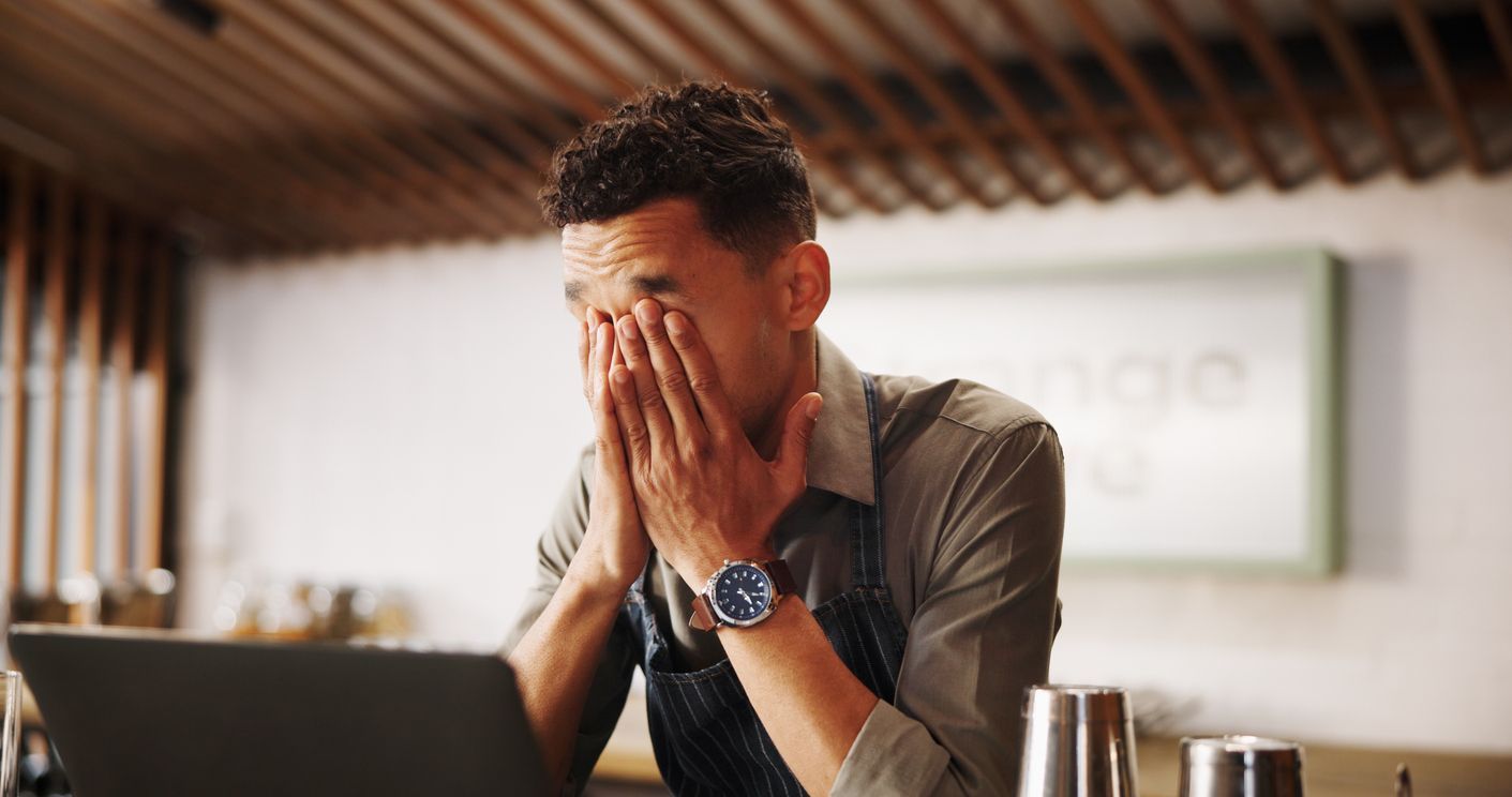 Man in apron at laptop, stressed and covering his face with hands.