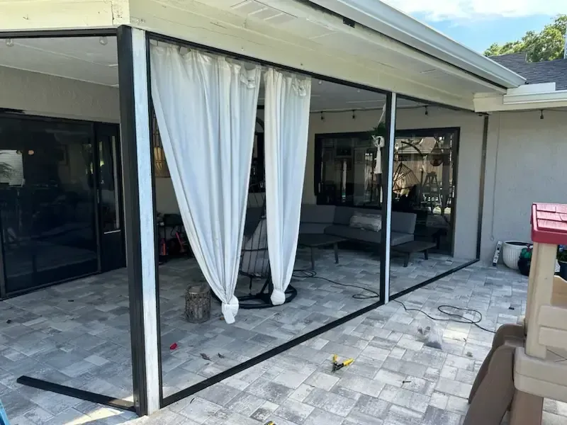 Covered patio with black-framed sliding doors, white curtains, and gray pavers overlooking an outdoor living area