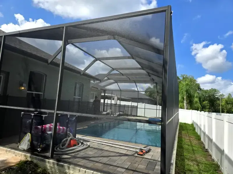 Screened-in backyard pool beside a gray house and white fence under a blue sky