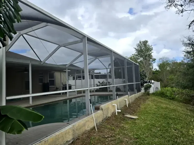 Screened-in backyard pool beside a gray house and white fence under a blue sky