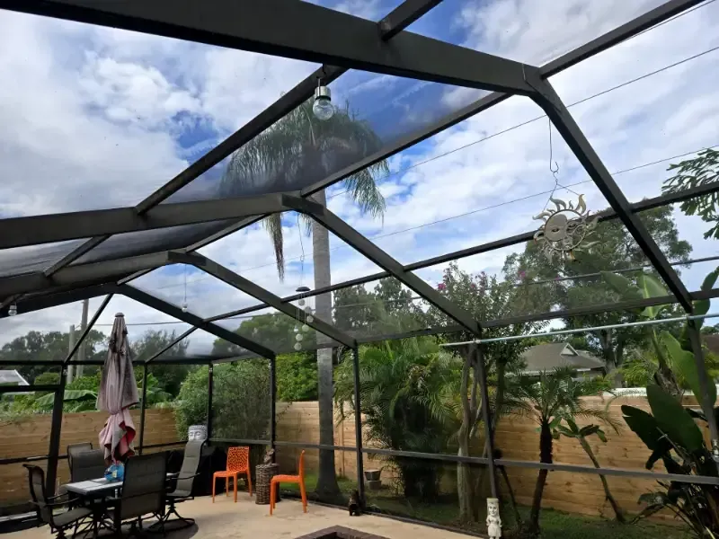 Screened patio with black metal roof beams, patio furniture, umbrella, and lush backyard greenery under a cloudy sky