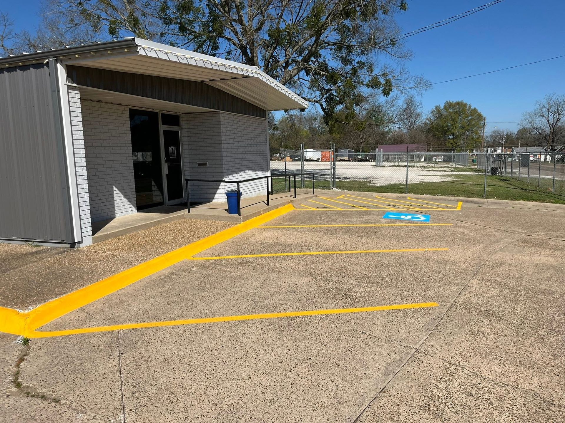 Small building beside a gravel lot with yellow painted lines and a blue bin under a clear sky