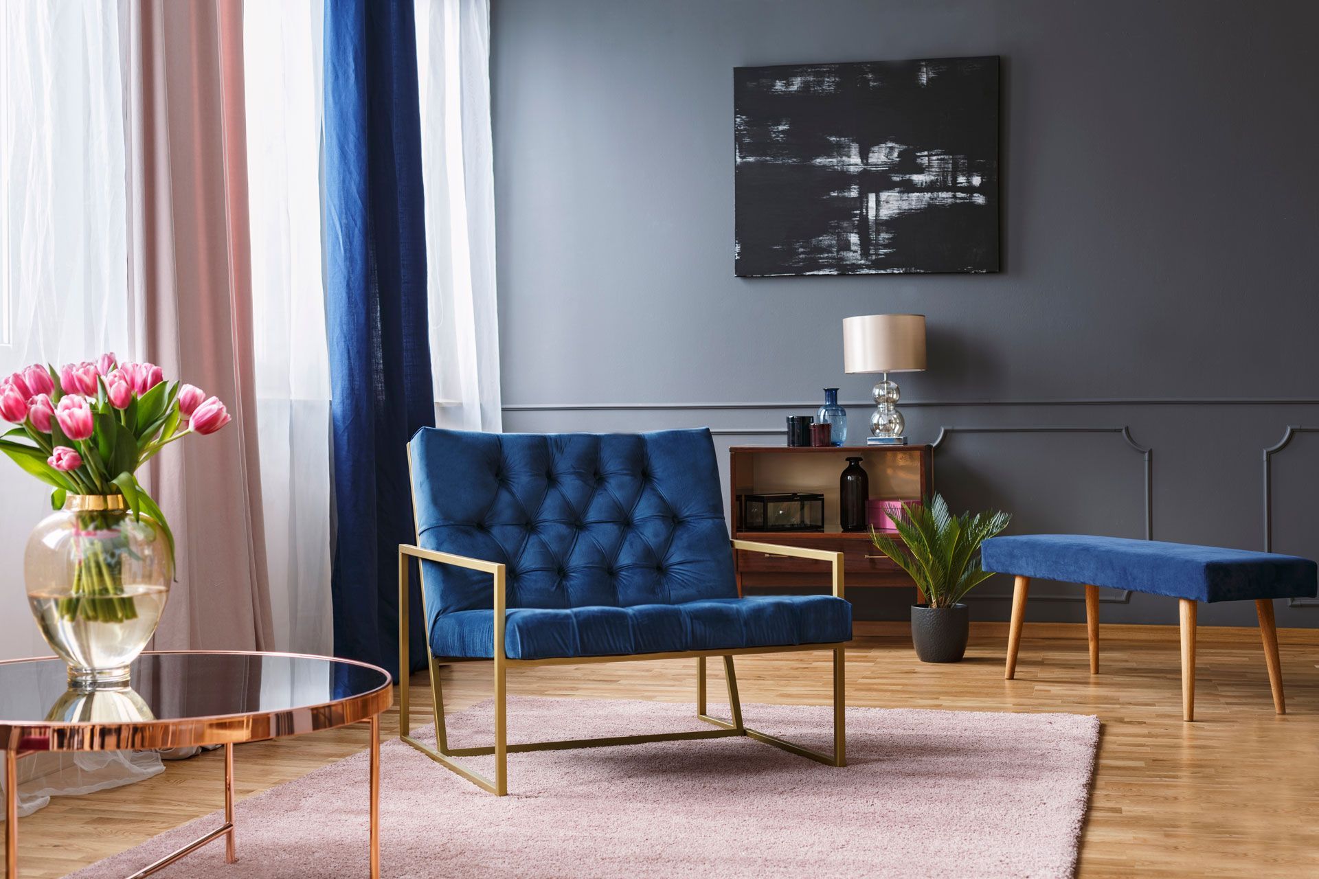 Blue velvet chair and bench in a gray room with pink rug, tulips, and abstract art.