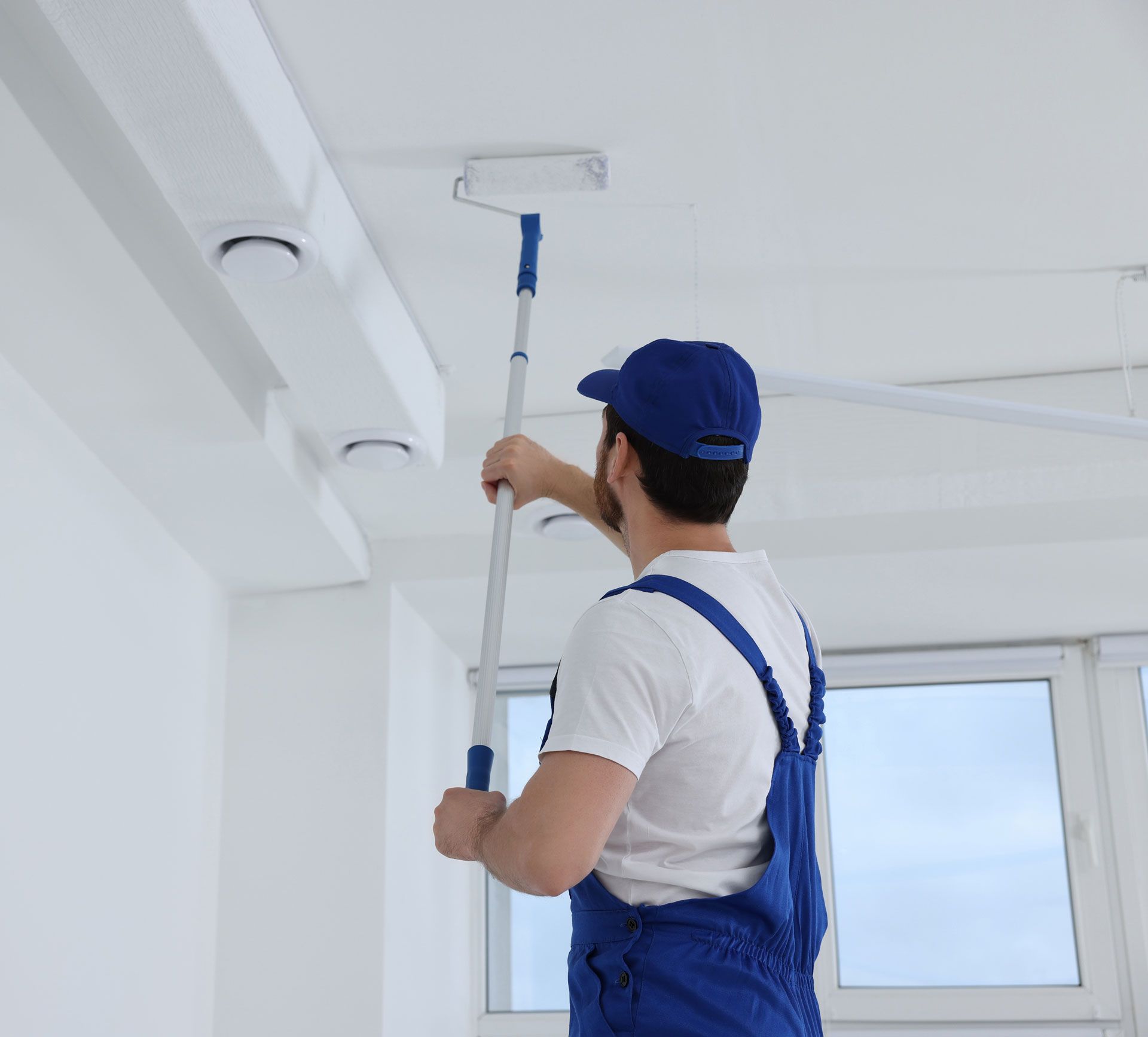 Painter in blue overalls and cap using a roller to paint a white ceiling.