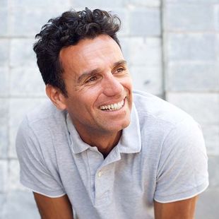Smiling man in a light gray polo shirt leaning outdoors against a pale wall