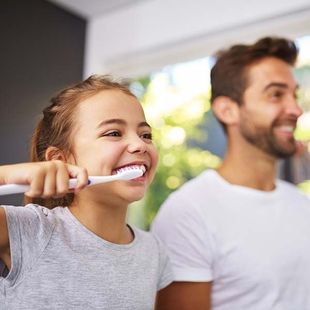 Child brushing teeth beside smiling adult in a bright bathroom