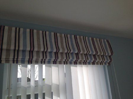 A striped roman shade is hanging over a window with vertical blinds.
