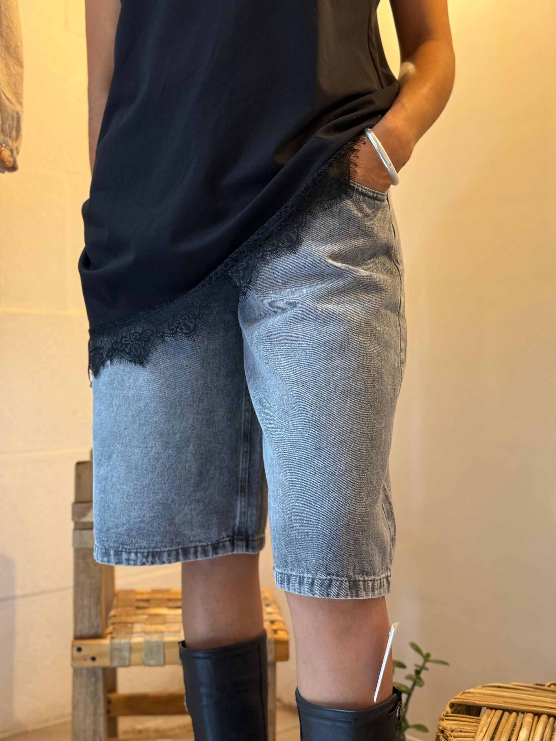 Persona de pie en el interior de un edificio, vestida con una camiseta azul marino, pantalones cortos vaqueros y botas negras, con la mano en el bolsillo.