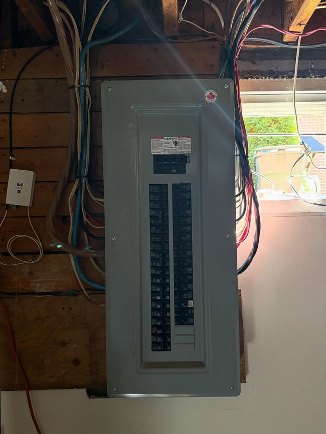 Un tableau électrique gris, fixé sur un mur à lattes de bois, dans une pièce avec une fenêtre à droite.