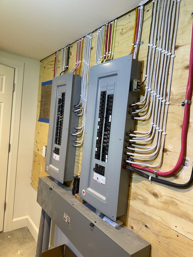 Deux panneaux de disjoncteurs électriques fixés sur un mur en contreplaqué, avec un câblage organisé remontant jusqu'au plafond.