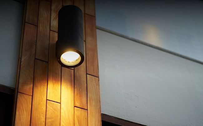 Une applique murale cylindrique noire, fixée sur une planche de bois verticale, projetant une lumière chaude vers le bas sur le bois.
