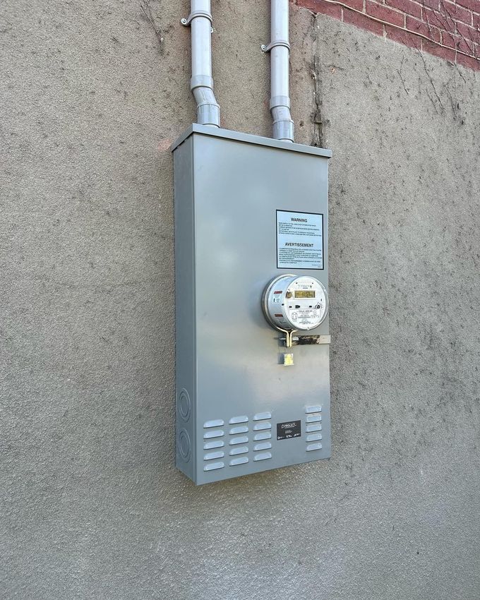 Un boîtier de compteur électrique gris fixé sur un mur en stuc, avec deux conduits s'élevant vers le haut.