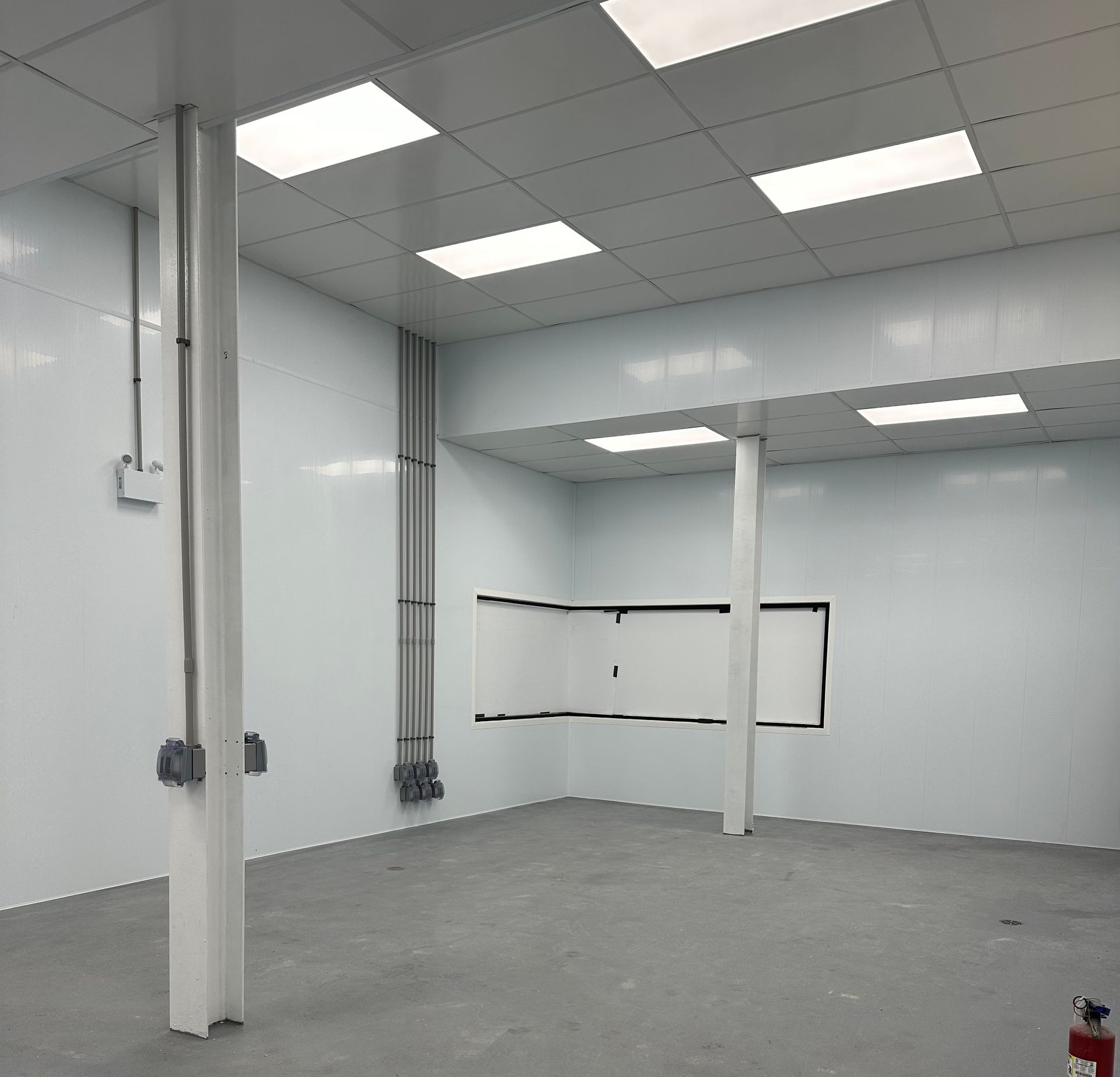 An empty, sterile-looking room with white panel walls, a grey concrete floor, white ceiling tiles, and two support poles.