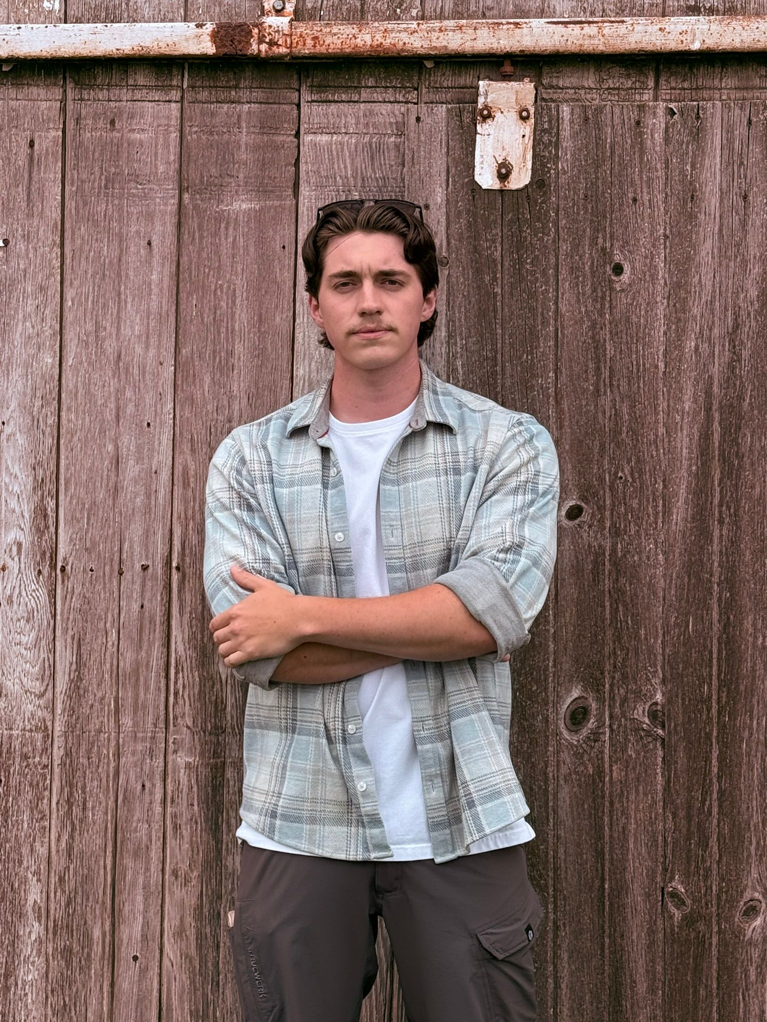 Man standing with arms crossed in front of a weathered wooden fence, wearing a light blue shirt.