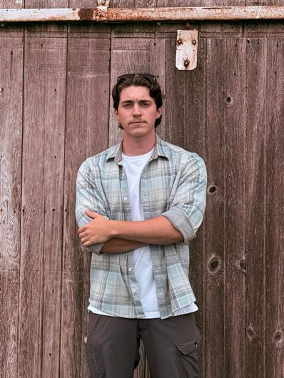 Man standing with arms crossed in front of a weathered wooden fence, wearing a light blue shirt.