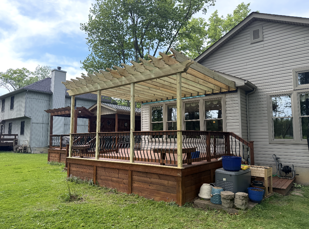 Backyard deck with wooden pergola attached to a house, surrounded by trees and outdoor items