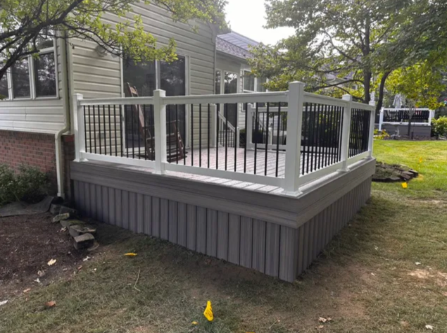 Gray elevated porch with white railings and steps beside a house, surrounded by grass and trees