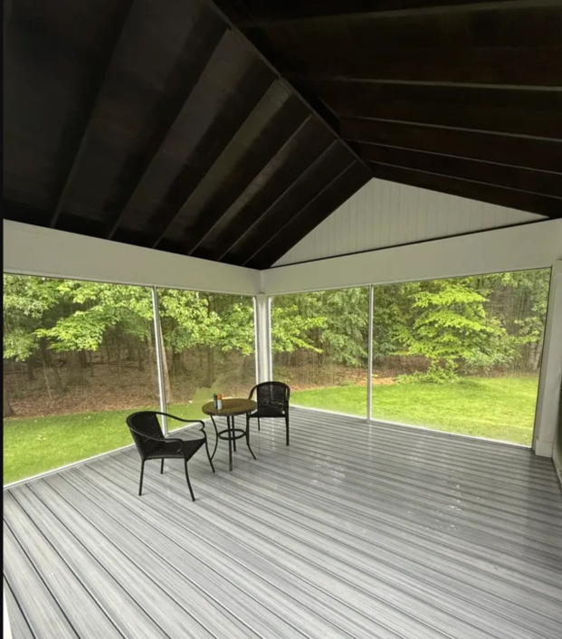 Covered deck with gray floor and dark roof, two chairs and a small table, overlooking a green yard.