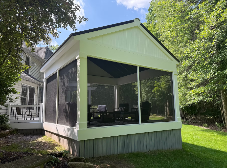 Screened porch addition with white trim and gray base beside a house, surrounded by trees and grass.