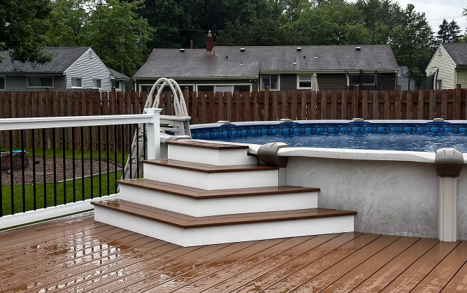 Backyard above-ground pool with white steps and railing on a wet wooden deck, fenced yard behind.