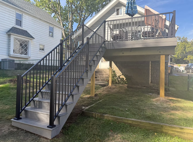 Raised wooden deck with black stairs and railing beside a house in a sunny yard