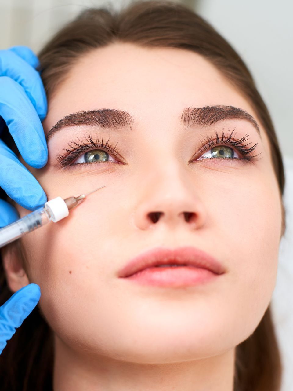 Woman receiving under-eye injections. Gloved hands hold a syringe near her eye. She looks upward with a neutral expression.