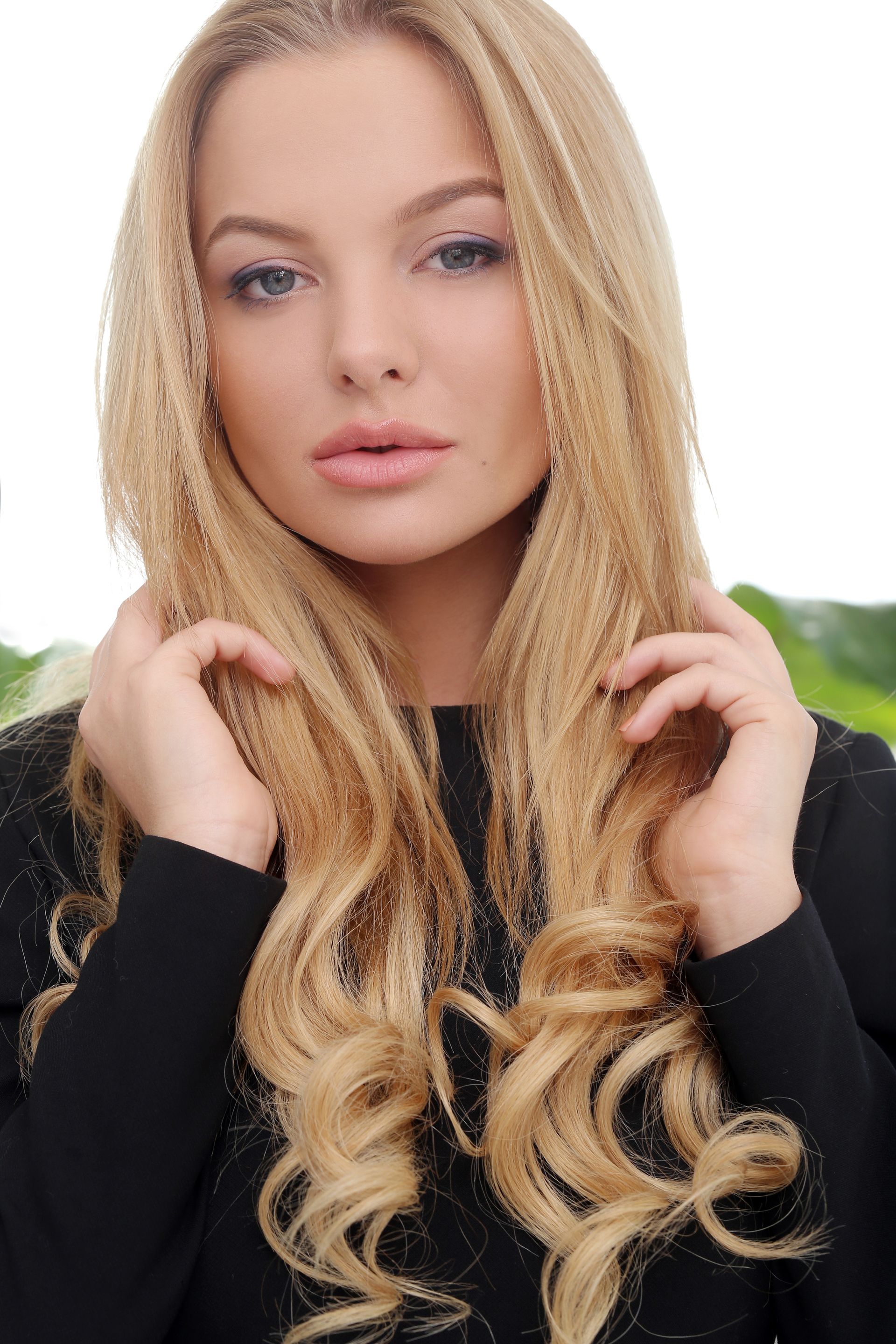 Blonde woman with blue eyes, wearing black, curls hair near her face. Outdoors, bright.