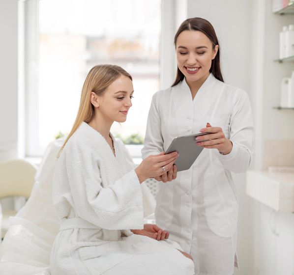 A cosmetologist showing a client a tablet in a bright, white spa. Both women are smiling.