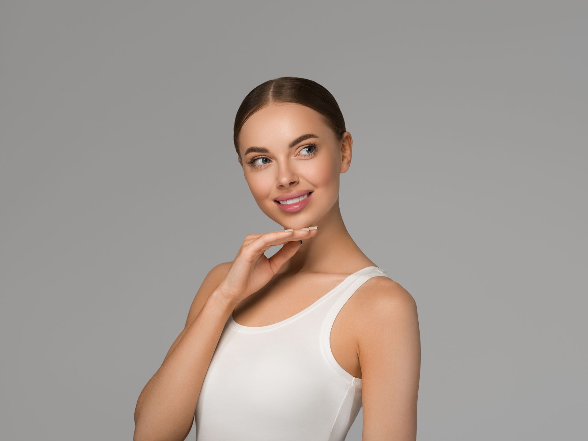 Woman in a white tank top, smiling and touching her chin, looking upwards; gray background.
