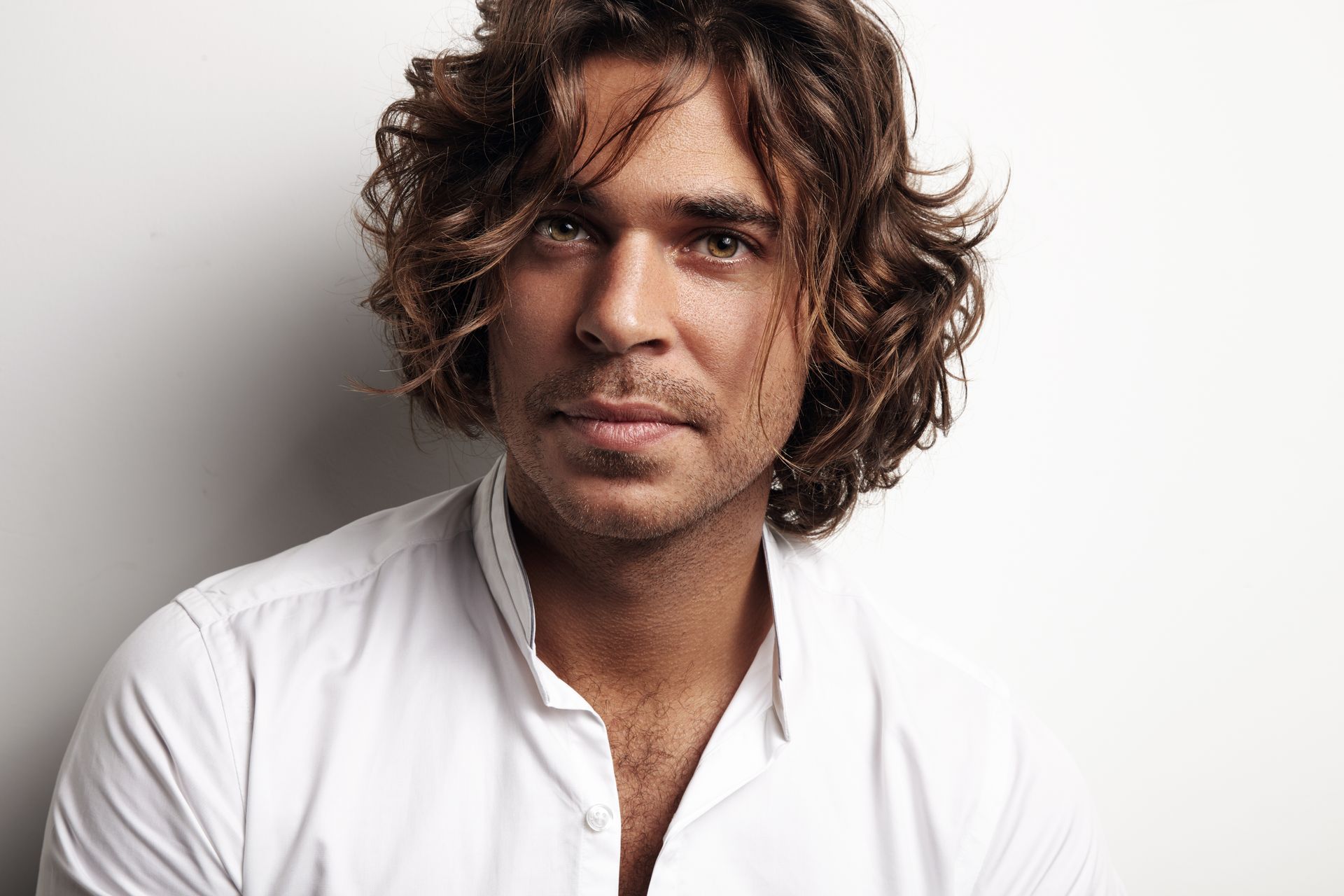 Man with long, wavy brown hair and a white shirt, looking directly at the camera against a white background.