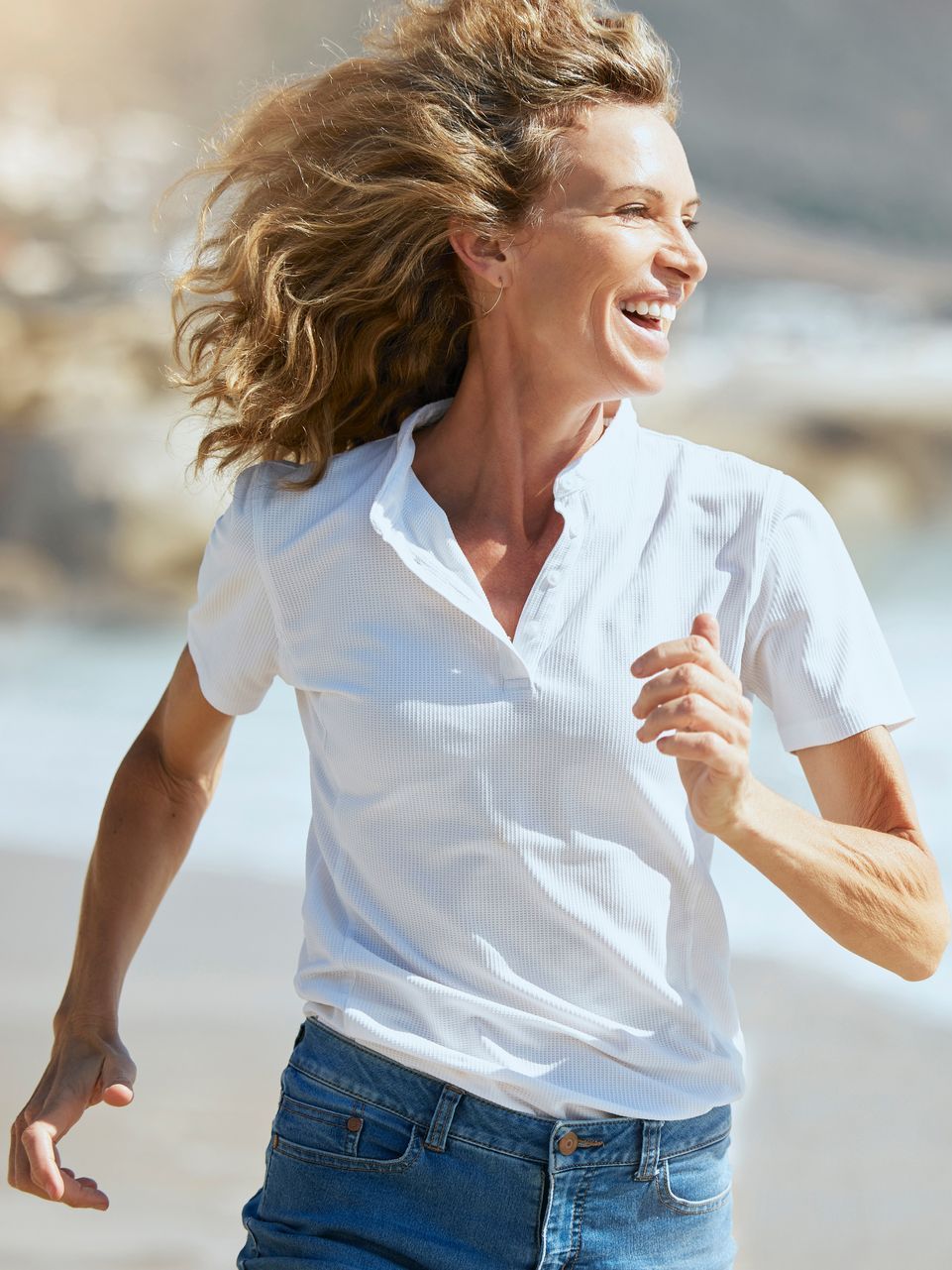 Woman with blonde hair running, smiling, on a beach wearing a white shirt and blue jeans.