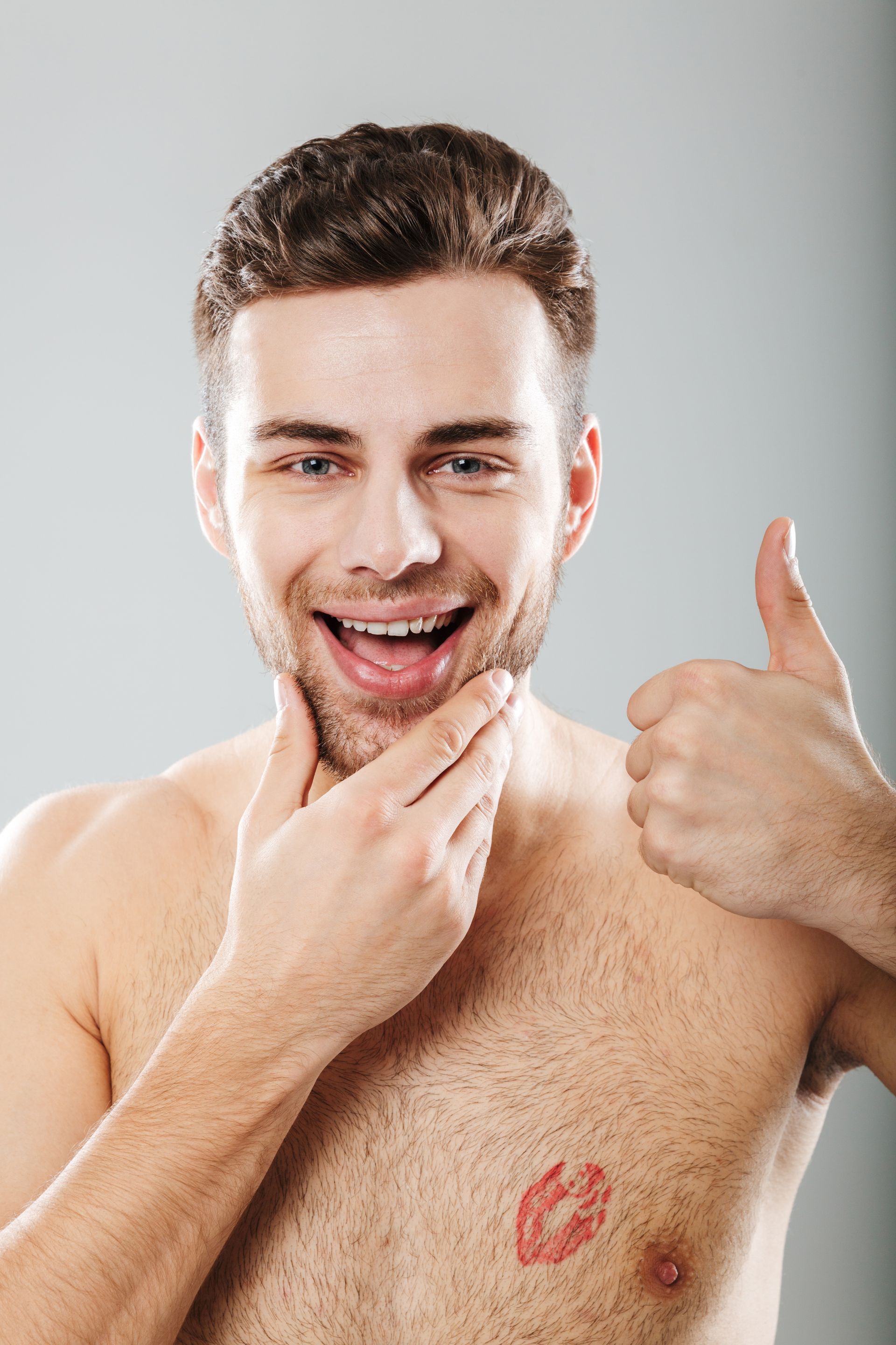 Man with lipstick kiss mark on chest, touching chin, giving thumbs up, smiling.