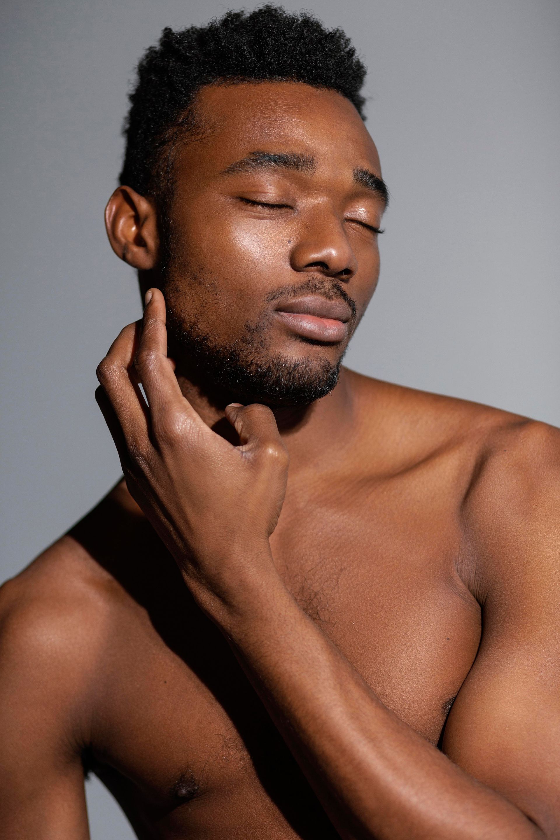 A Black man with closed eyes touches his neck, appearing serene. Against a gray backdrop, bare-chested.