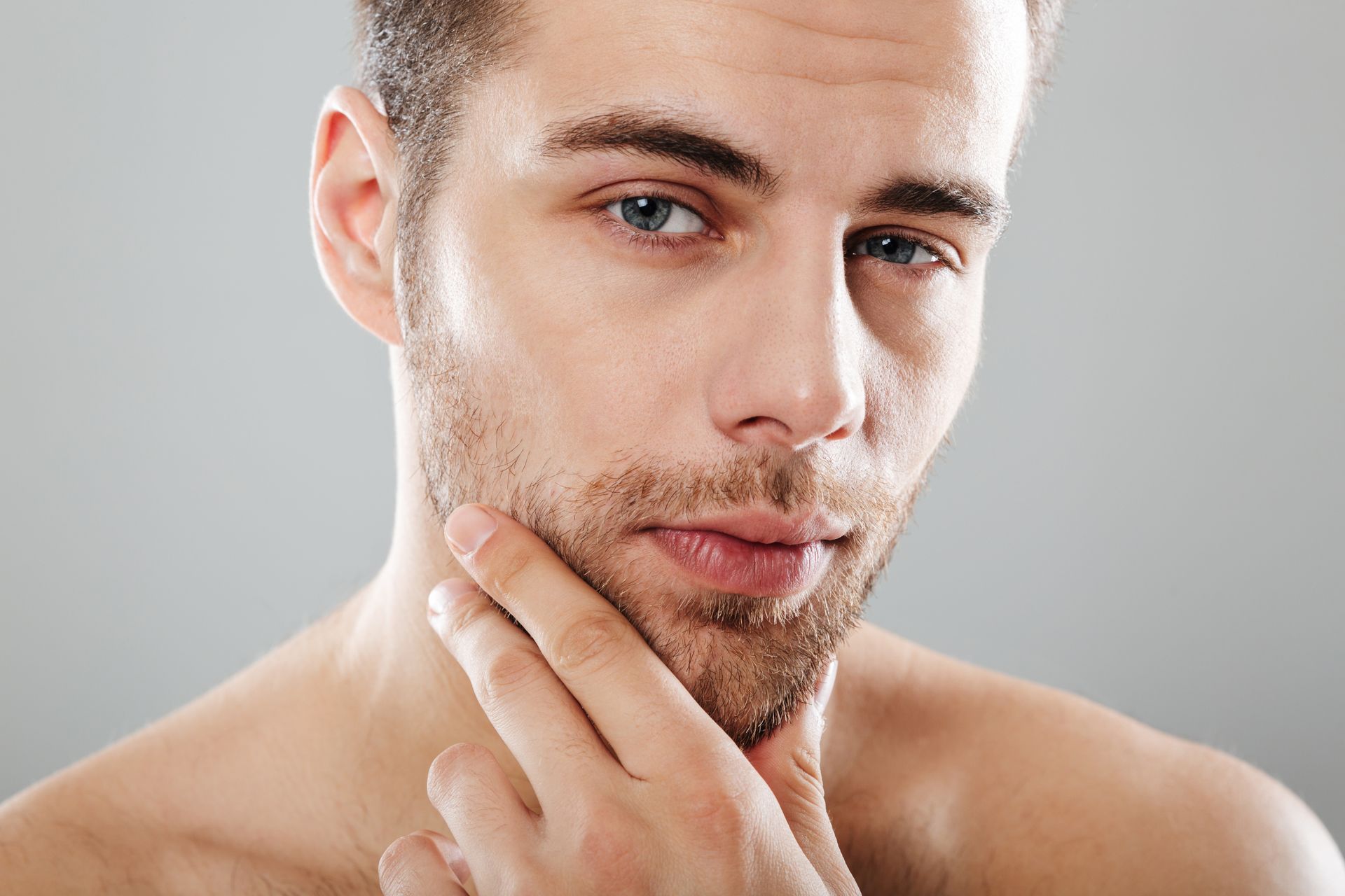 Man with short beard, gazing at viewer, touching his chin, against a gray backdrop.