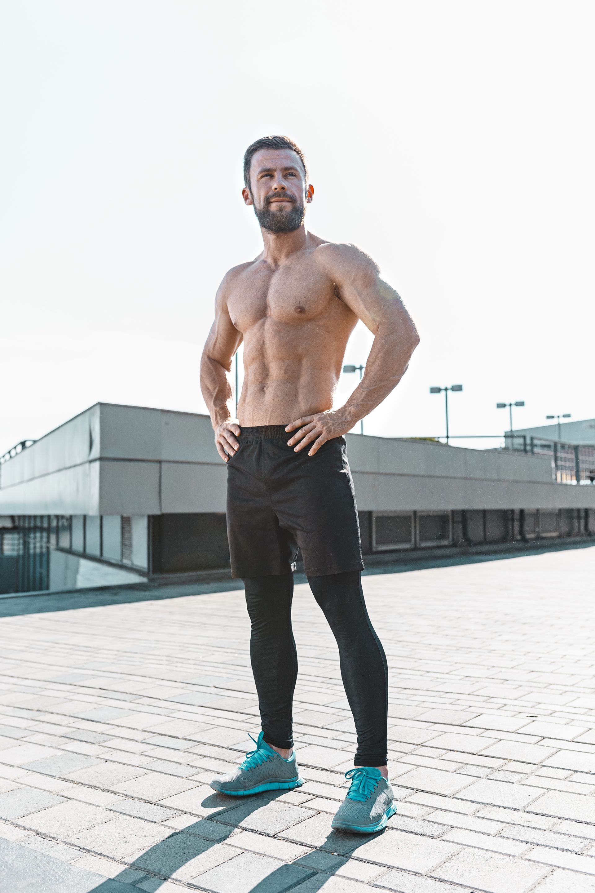 Fit, shirtless man standing on a rooftop, hands on hips, looking up; wearing black pants, sneakers.