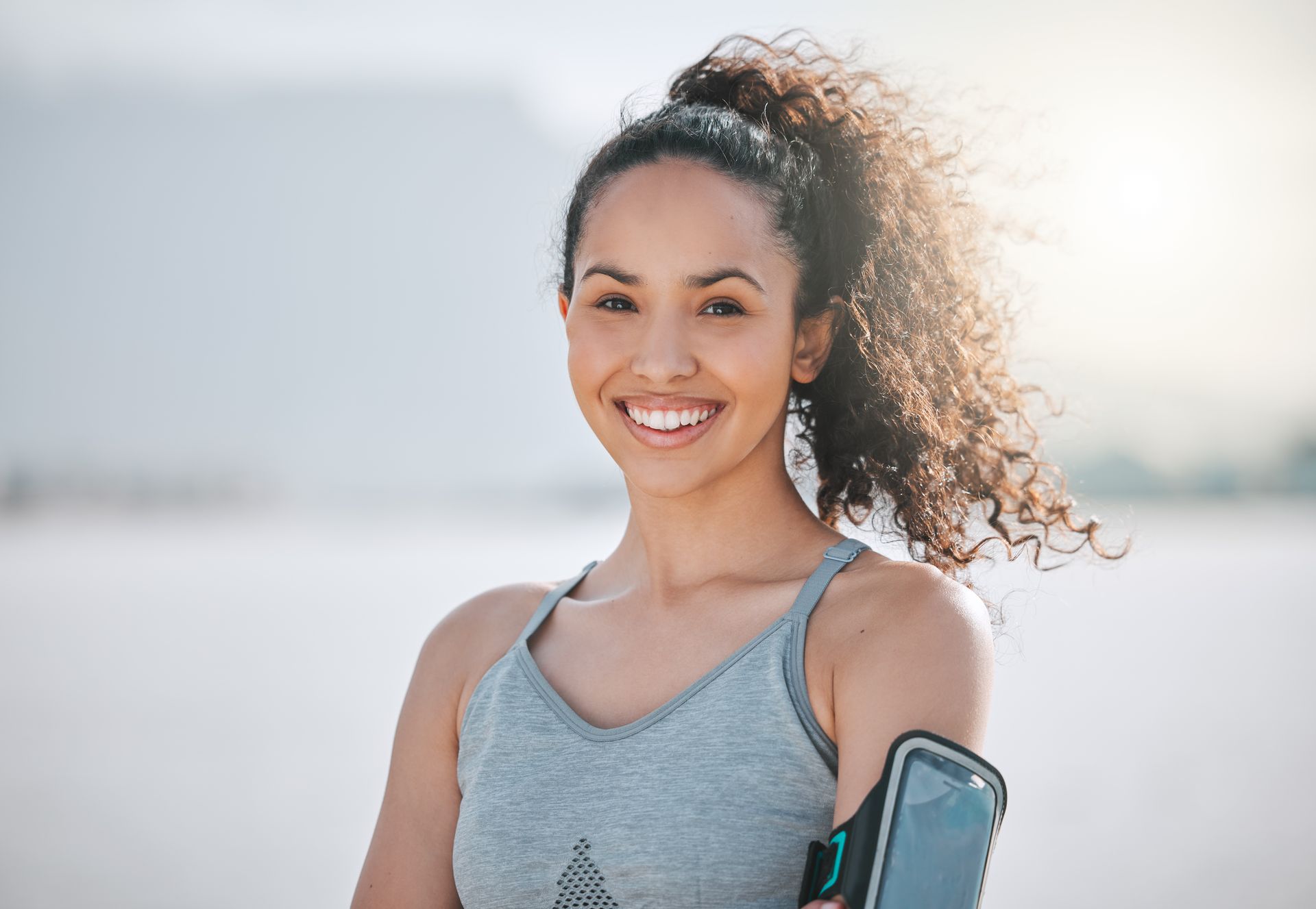 Woman with curly hair smiles, wearing athletic wear, arm with phone holder, outdoor setting.
