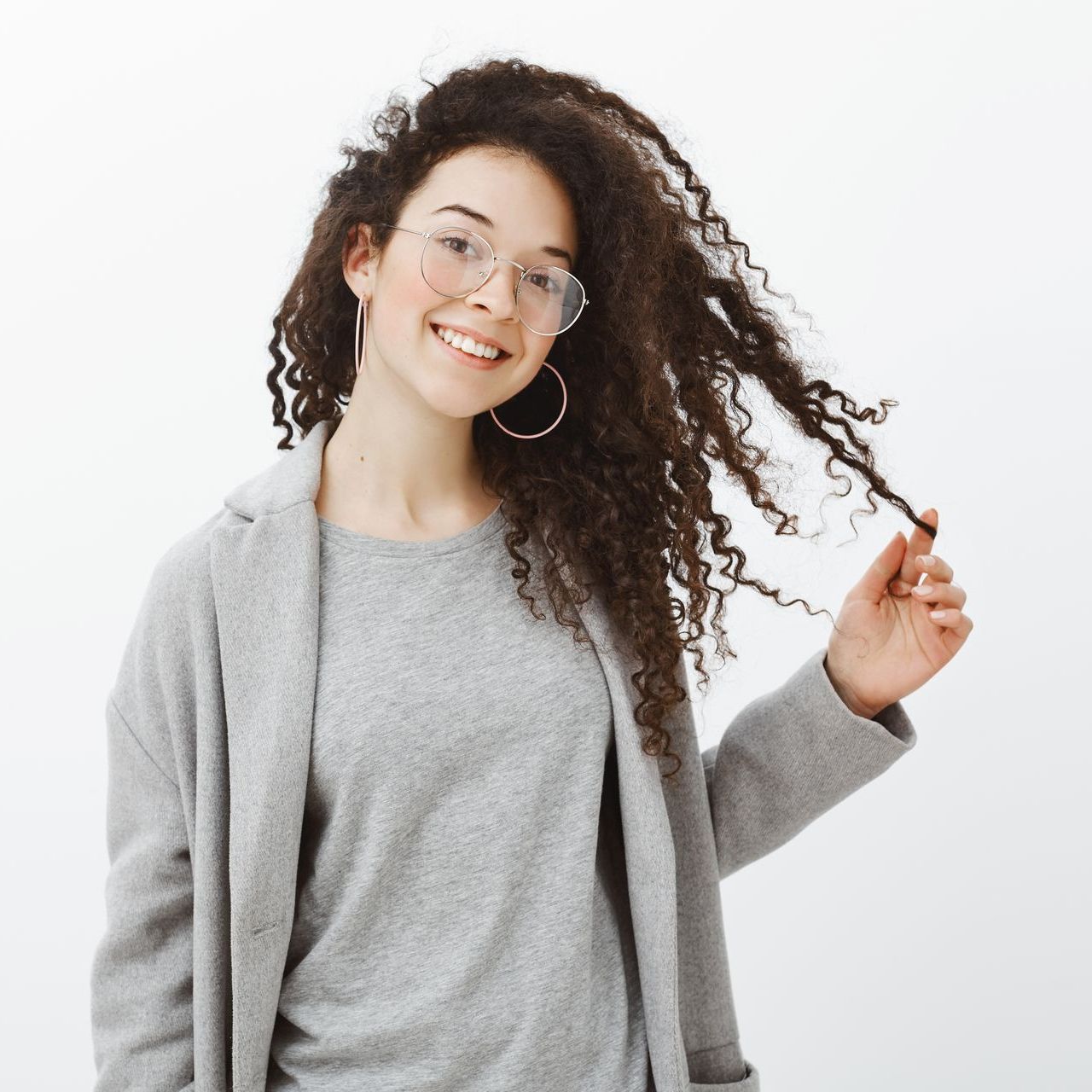 Woman with curly hair, glasses, smiling, holding hair, wearing gray sweater and coat.