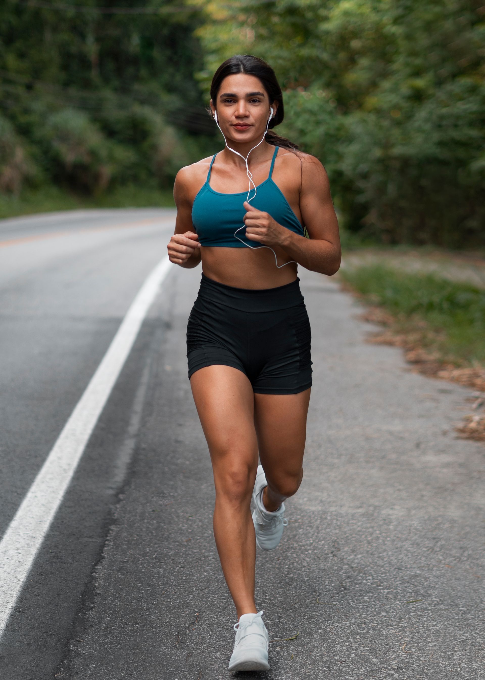 Woman running on a road wearing a teal sports bra, black shorts, and earphones; surrounded by trees.