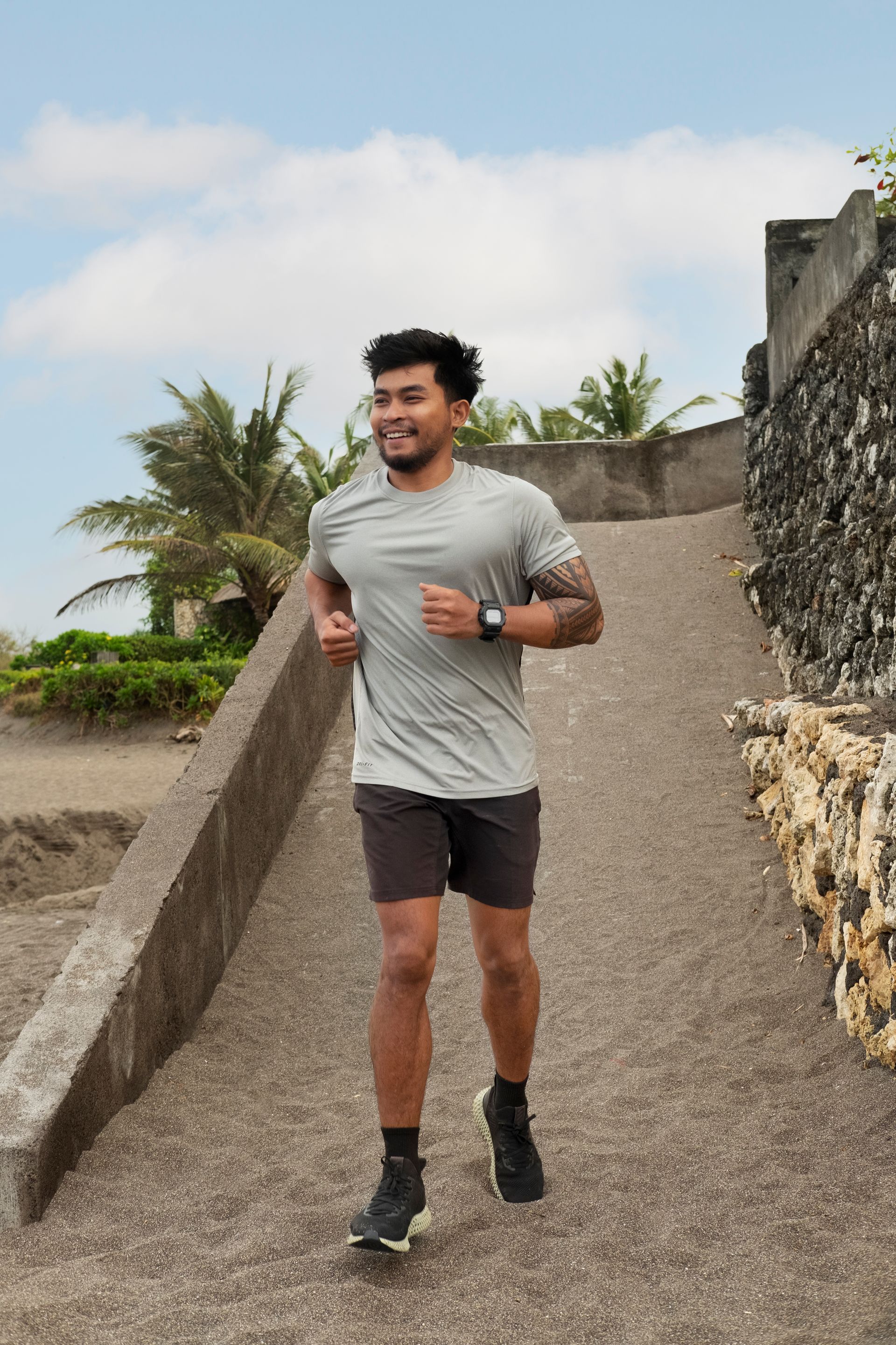 Man running up a gravel path, smiling. Wearing gray shirt, dark shorts and shoes. Outdoors with palm trees and stone wall.