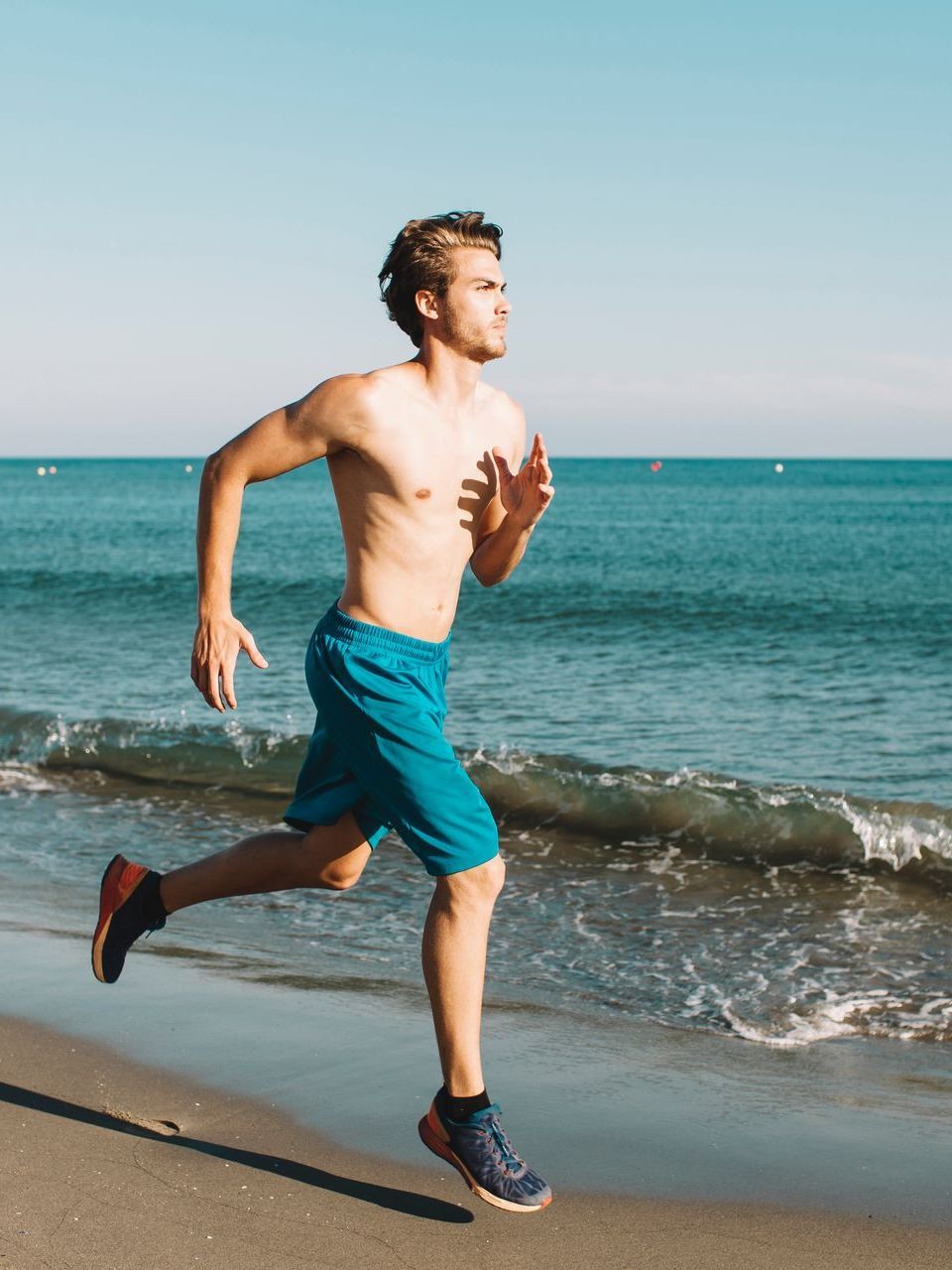 Man running shirtless on beach, blue shorts and shoes, ocean in background.