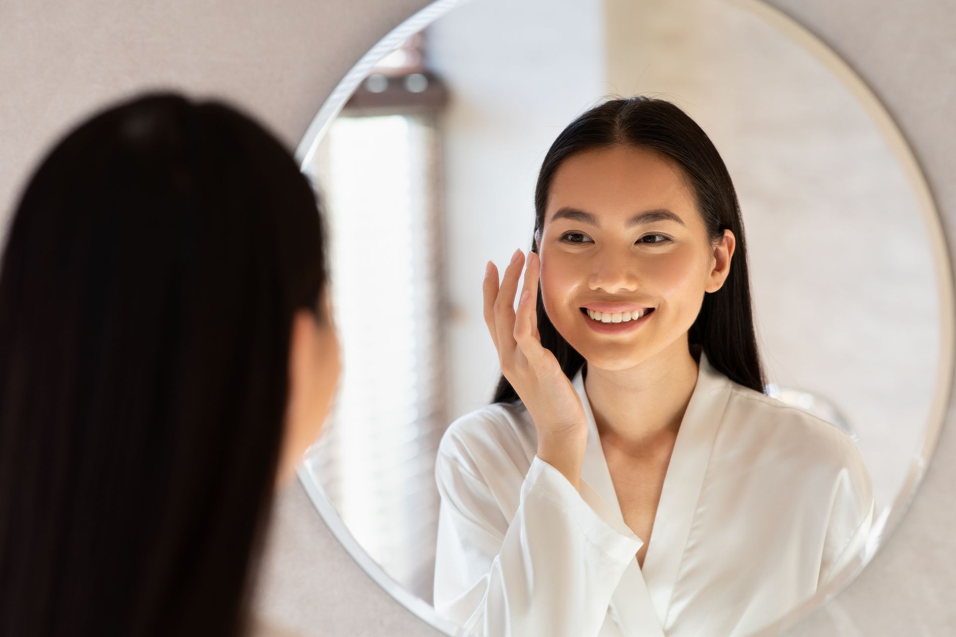 Woman in a white robe smiles, looking at her face in a circular mirror, indoors.