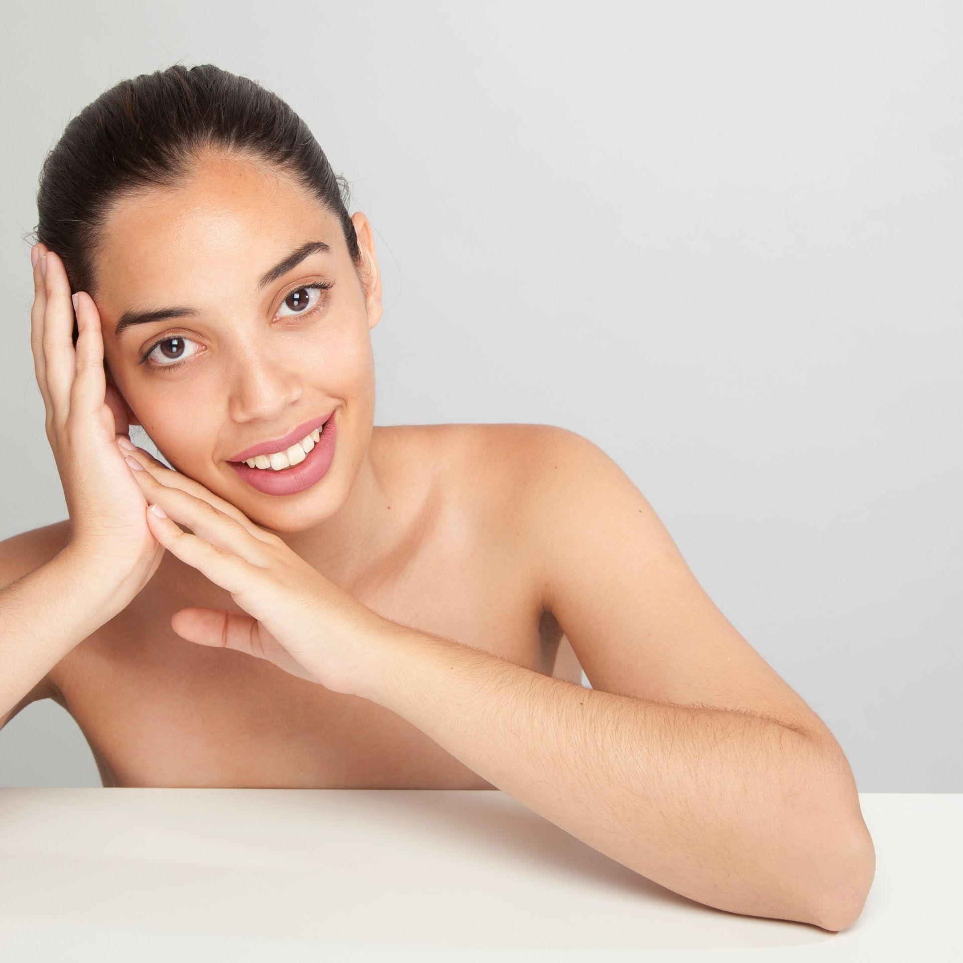 Woman with brown hair, smiling, resting face on hands, leaning on white surface.
