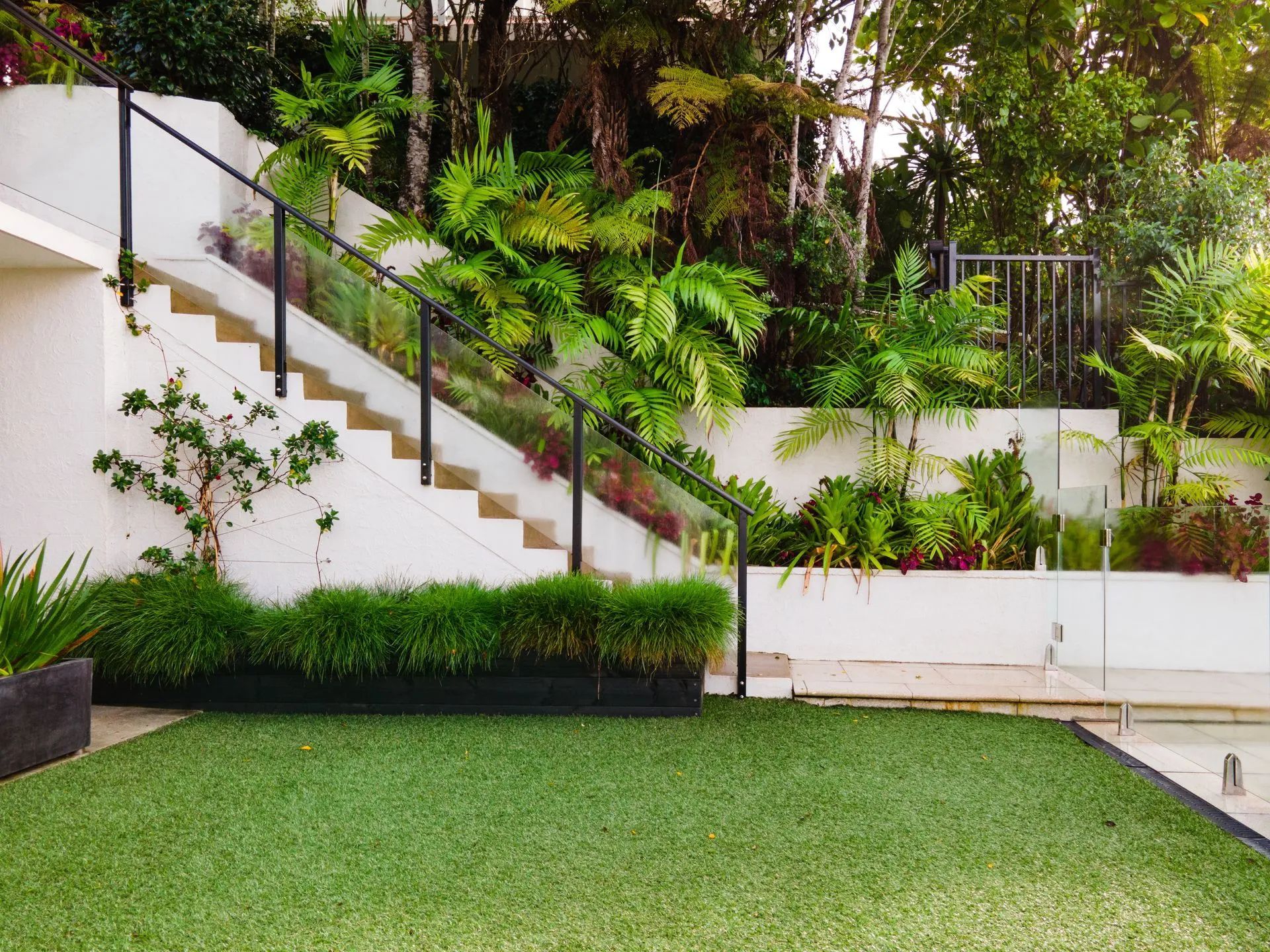 White staircase with black railing and greenery, ascending a hillside garden with tropical plants.