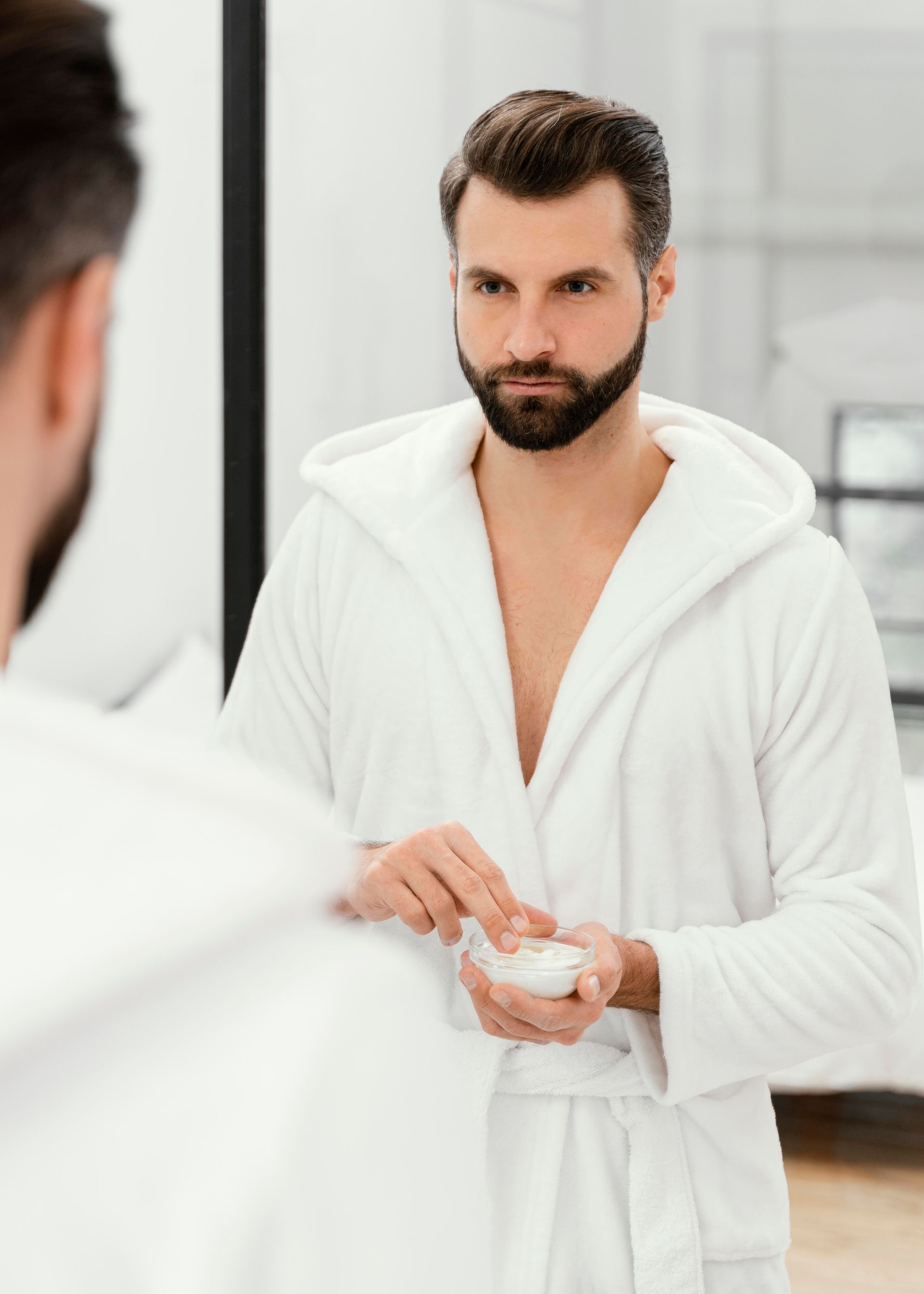 Man in white robe looking in mirror, holding cream.