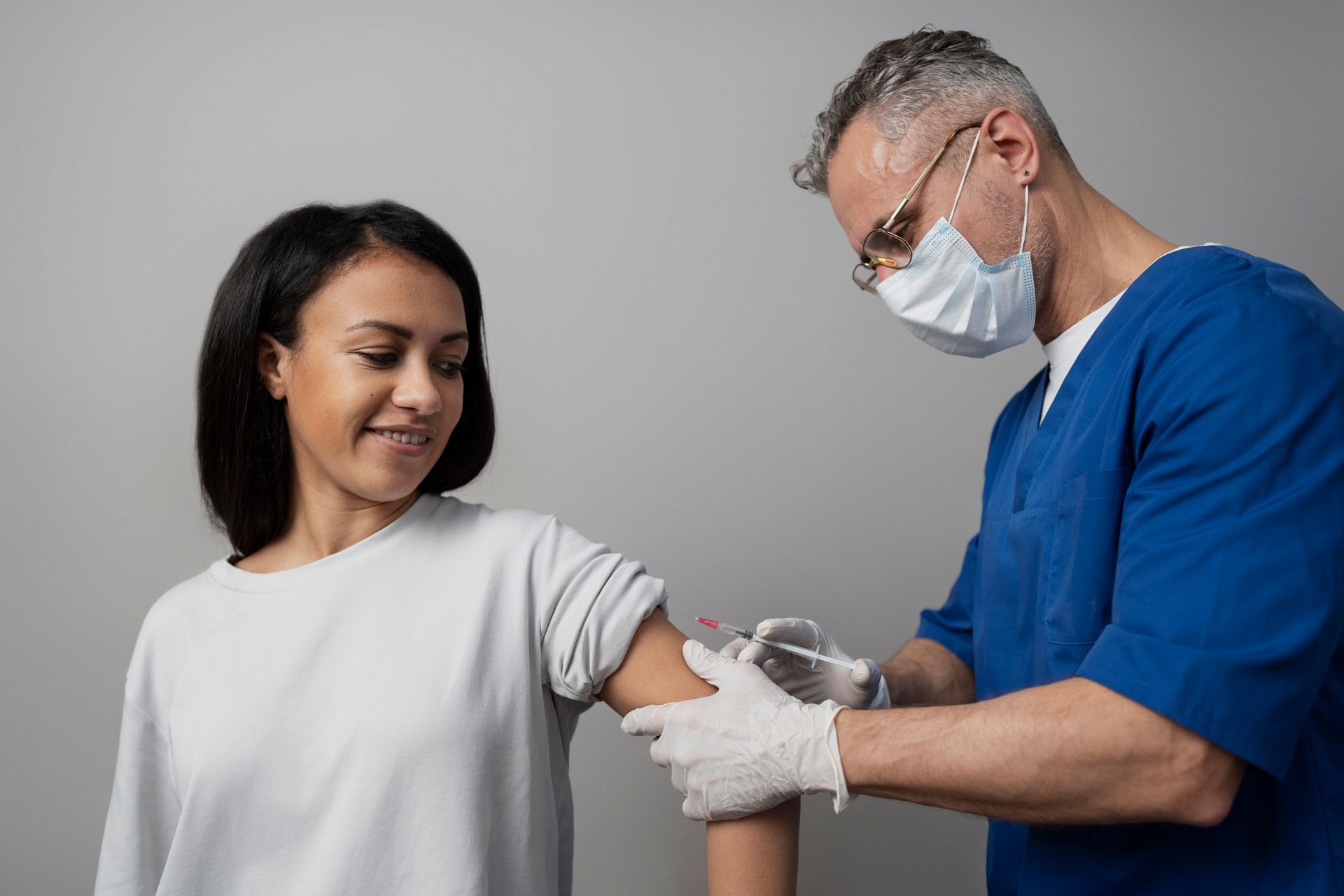 A medical professional gives a Black woman a shot in the arm against a gray background.