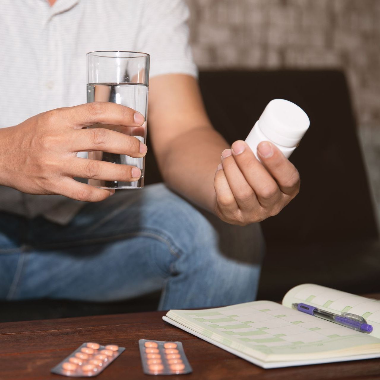 Man taking pills with a glass of water, sitting on a couch with a notebook and pills.