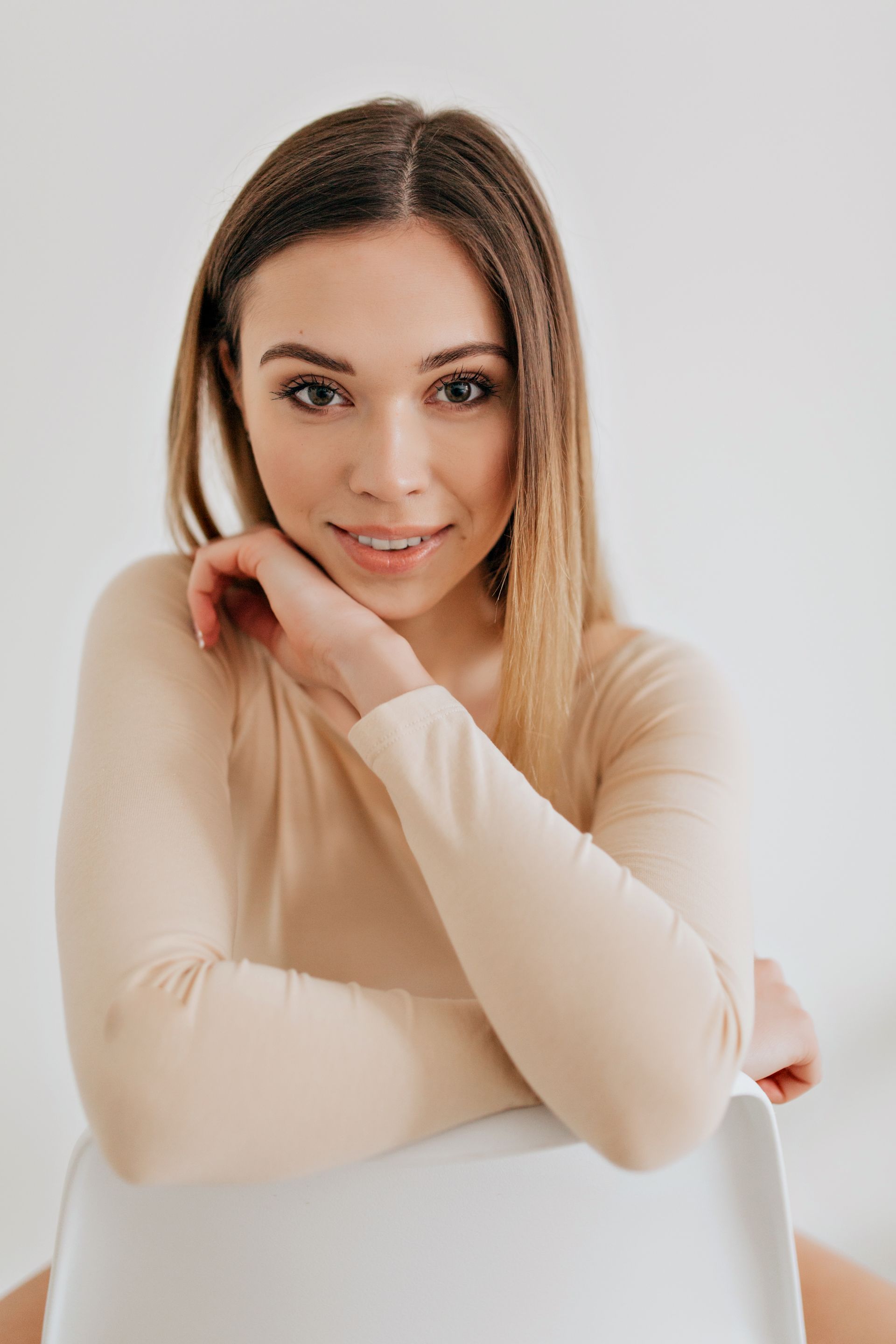 Woman with long hair, smiling, leaning on a white chair, wearing a beige bodysuit.