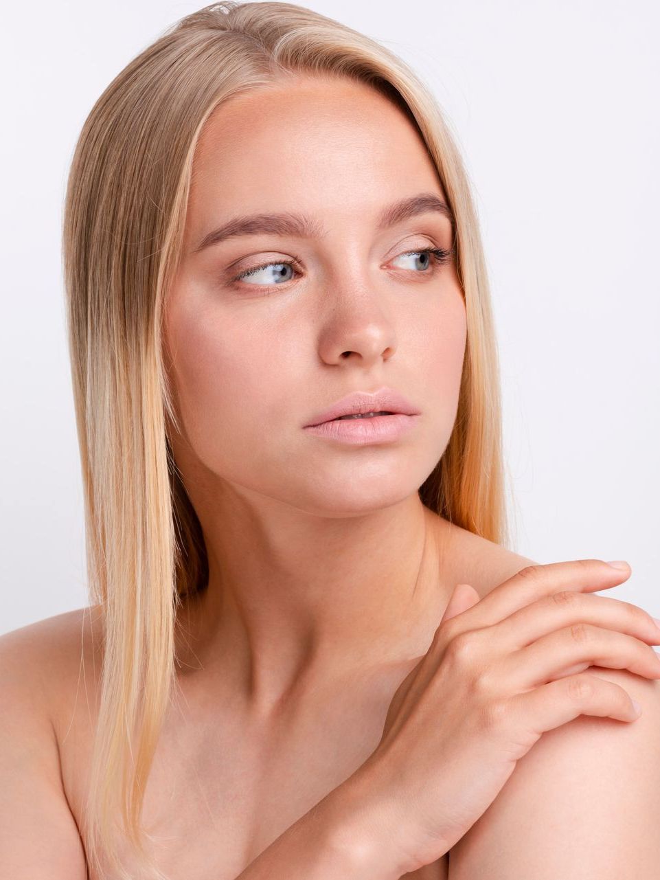 Blonde woman with fair skin looking off to the side, hand on her shoulder, on a white background.